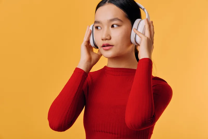 Woman in red sweater wearing headphones standing against orange background looking sideways with thoughtful expression and hands adjusting modern wireless headset.