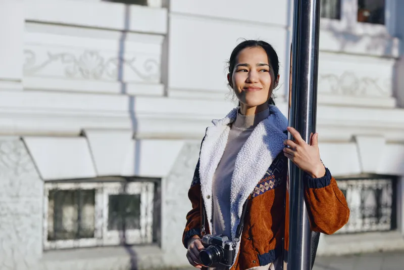 Woman with a camera outdoors beside a pole, urban backdrop, fashionable coat and scarf, natural light, candid street style perfect for fashion and travel stock