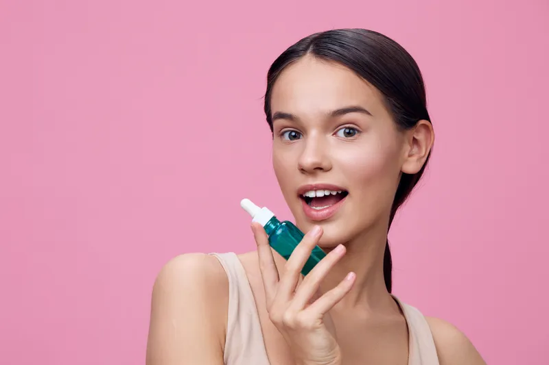 Young woman holding a green serum, smiling against a pink background, showcasing skincare and beauty concept emphasizing healthy skin and self care