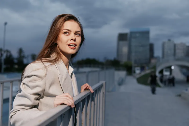 Young woman in beige blazer outdoors in urban environment with confident smile and modern cityscape background at dusk.