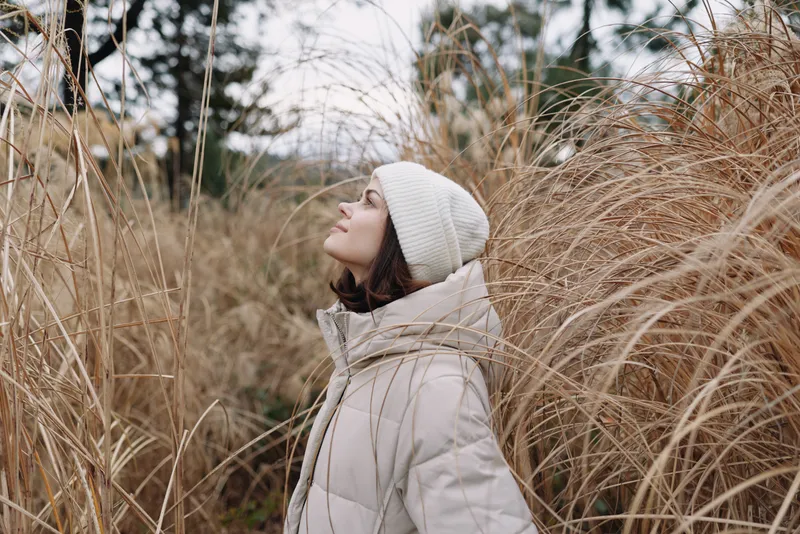 Woman in a light puffer coat and beanie gazes upward among tall grasses, capturing calm outdoor winter nature and quiet contemplation, peaceful mood.