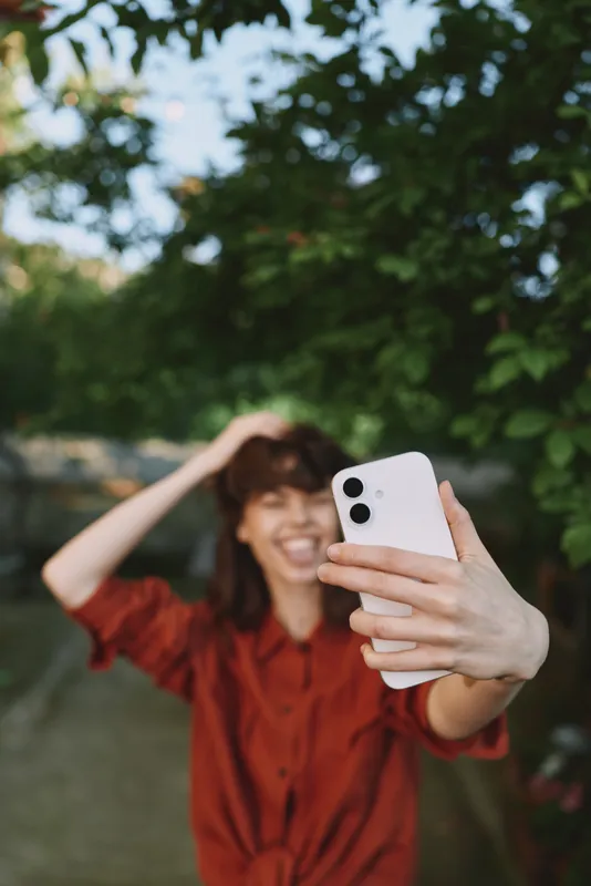 A smiling woman in casual red top takes a selfie outdoors in a sunny park this summer, smartphone visible, capturing a portrait lifestyle moment for everyday fashion
