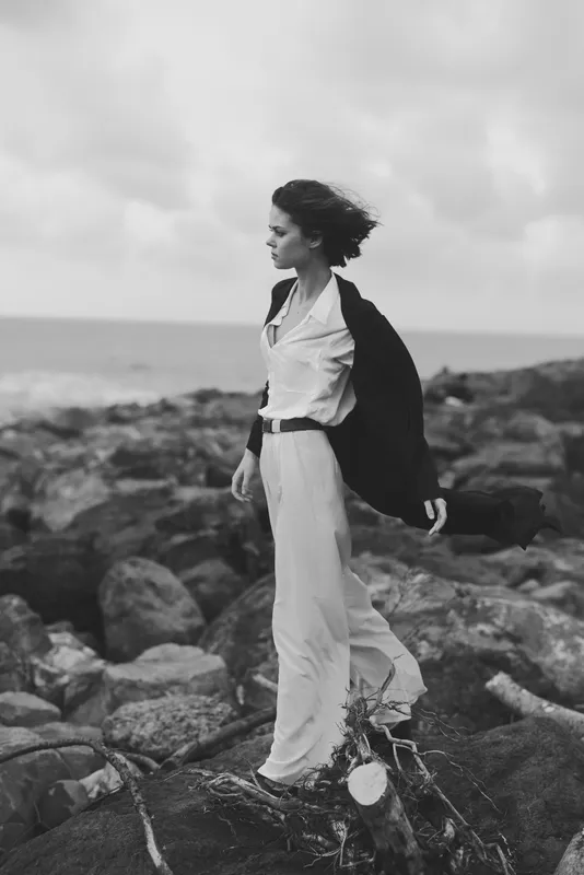 Woman stands on a rocky shore, dress flowing in the wind, coat draped over her shoulders, gazing toward the sea in a moody monochrome scene