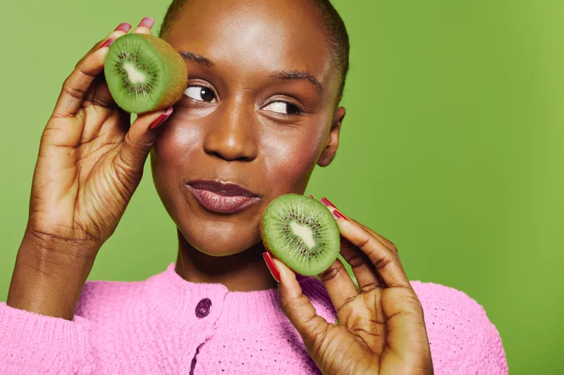 Young black woman with dark skin holds two halves of fresh kiwi near her face. Studio portrait on green background. Concept of healthy lifestyle, fruit, and natural beauty.