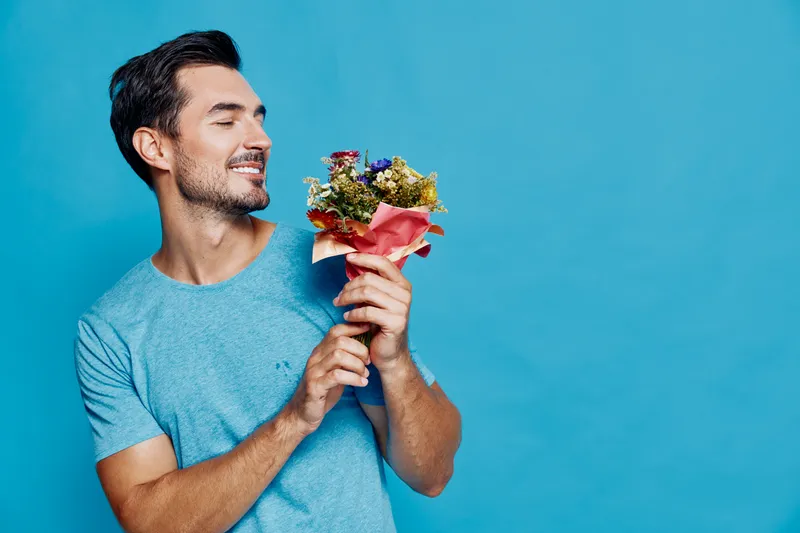 Happy young man smiling while holding a bouquet of colorful flowers against a vibrant blue background. Joyful emotions and cheerful mood captured in this portrait.