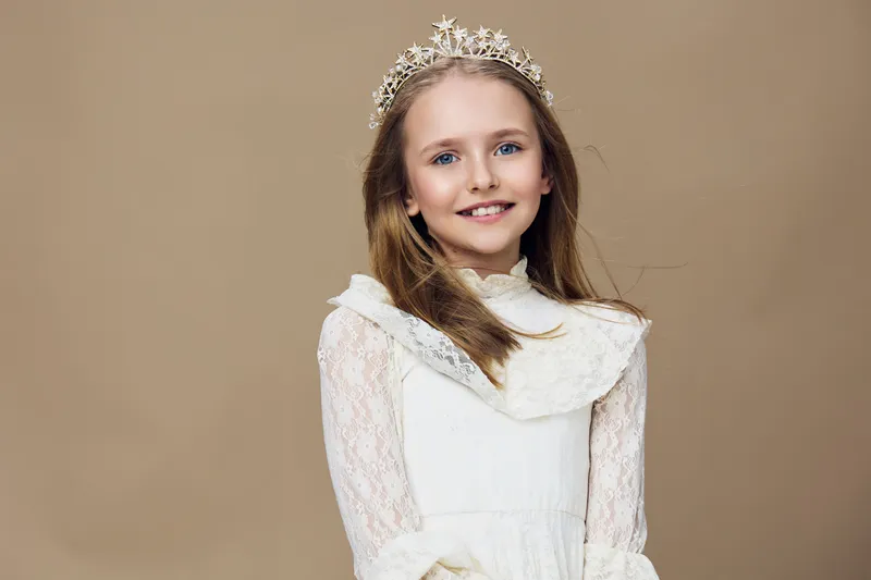 Portrait of a young girl in a white dress with a crown, conveying innocence and joy against a warm beige background, perfect for seasonal celebrations