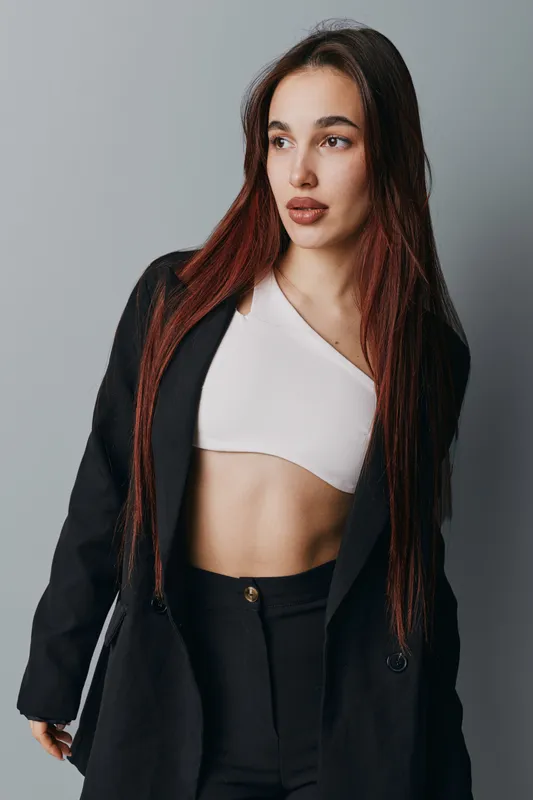 Woman with long hair wearing black jacket and white crop top posing against gray background looking sideways with confident expression in studio portrait fashion style.