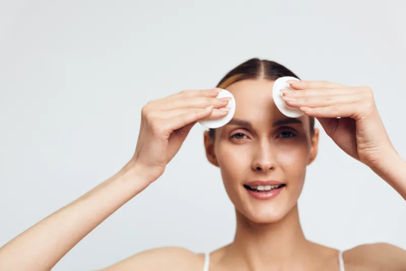 Young woman with clear skin removing makeup with cotton pads on white background. Concept of skincare, cleansing, beauty routine, and healthy lifestyle. Soft lighting highlights natural look.
