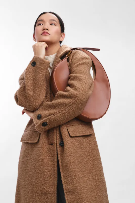 asian woman wearing a tan wool coat and round handbag stands against a simple backdrop, striking a thoughtful pose with chin resting on hand, conveying modern fashion and confident elegance.