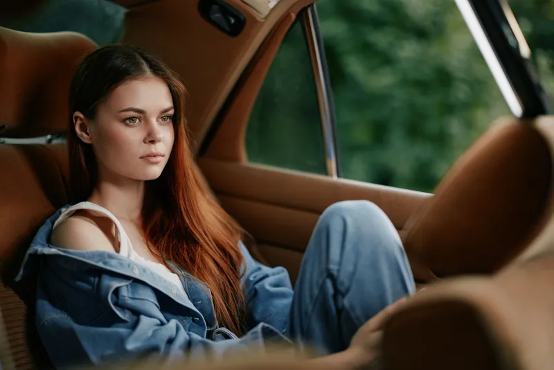 Young woman with long hair in casual denim outfit sitting pensively in a vintage car, surrounded by nature, conveying a sense of solitude and reflection.