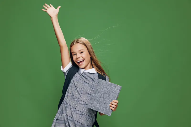 Girl student smiling and raising hand with backpack and tablet in hand for education, learning and school success in front of green background.