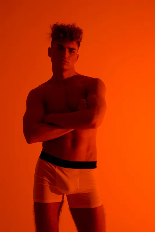 Young man with muscular body and curly hair posing with crossed arms in boxer briefs against bright orange background, emotive and confident male fitness portrait.
