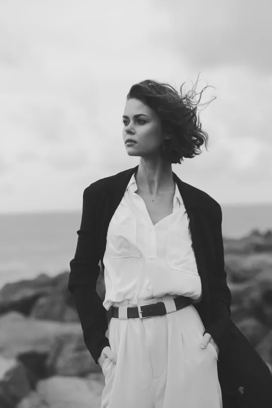 Stylish woman wearing a white suit and black coat stands confidently by a rocky coast, wind sweeping her hair, creating a bold monochrome fashion moment.