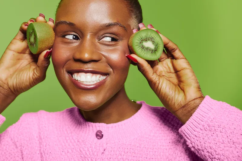 Smiling African American woman with dark skin holds two kiwi halves near face wearing pink sweater. Concept of healthy lifestyle, fresh fruit, wellness and natural beauty in studio.