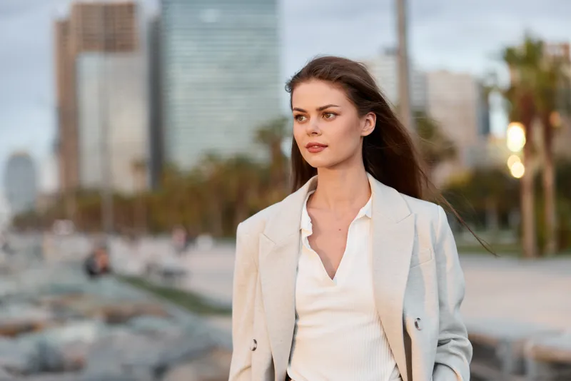 Young woman in light beige blazer and white top confidently walks urban seafront with casual hairstyle, natural makeup, calm expression, city skyline and palm trees background