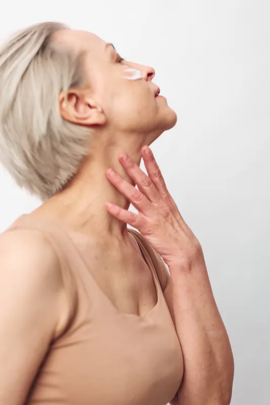 Mature woman with short blonde hair applies white eye patches while gently touching her neck. Concept for skincare, anti aging, self care, and beauty routine on neutral background.