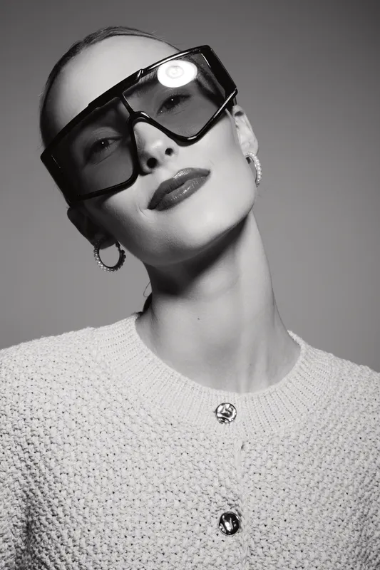 Stylish white woman with pale skin wearing oversized sunglasses and hoop earrings poses confidently in black and white studio portrait. Fashion and elegance concept.