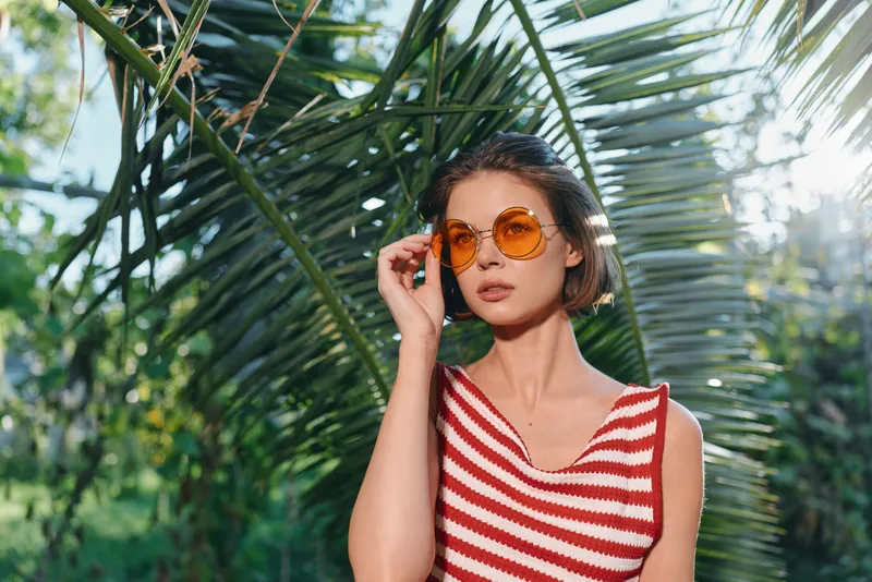 A stylish young woman wearing a red and white striped sleeveless top and round orange sunglasses stands outdoors among lush tropical plants, exuding a sunny, carefree fashion vibe.