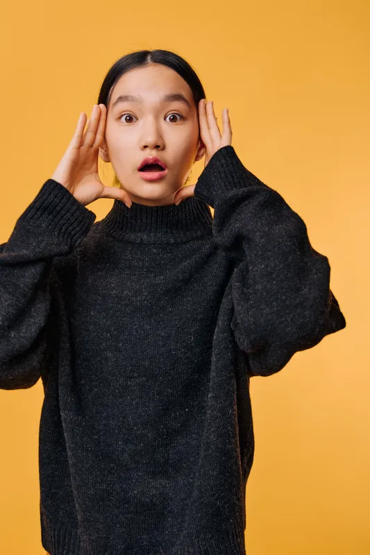 Young woman with black hair wearing dark sweater showing surprise with hands near face on yellow background. Female portrait with astonished expression and casual outfit in studio light.