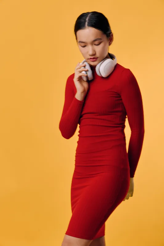 Woman wearing red dress and white headphones holds small object near face on yellow background. Beauty, casual style, technology, fashion and studio portrait with thoughtful mood and minimalism.