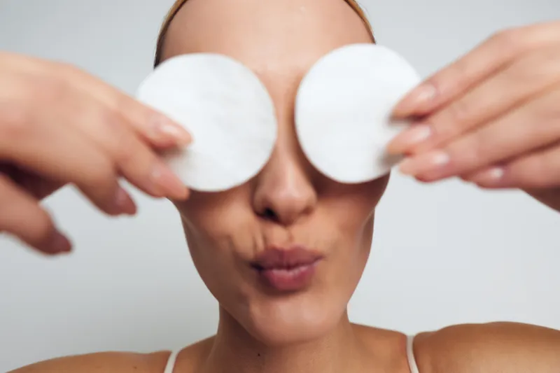 Young woman with bare shoulders holds two cotton pads over her eyes while puckering her lips against a neutral background expressing playful skincare routine mood.
