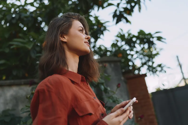A woman in a rust shirt stands outdoors, looking up with a phone in hand. A lifestyle moment capturing curiosity, calm, and everyday creativity in the photo
