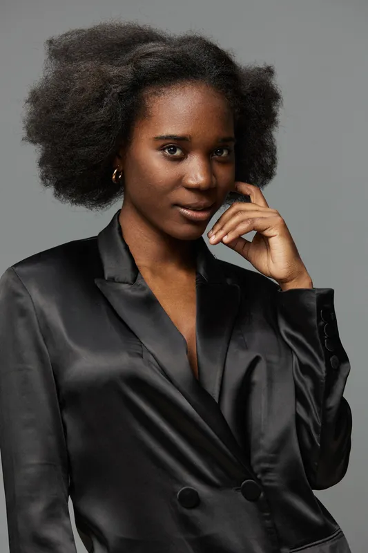 Confident young woman in a black satin blazer showcasing natural beauty and style against a neutral gray background, perfect for fashion and empowerment themes