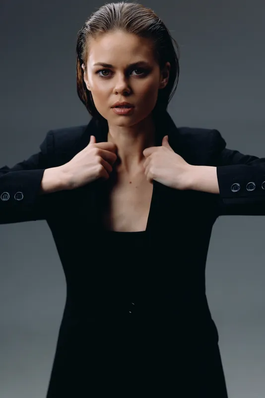 Serious woman in black outfit posing confidently with hands near collar against dark background.