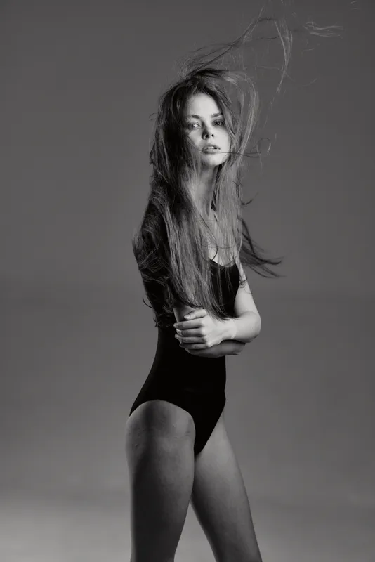 Black and white portrait of a young woman with long hair in a bodysuit, expressing confidence and elegance in a studio setting.