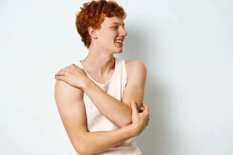 Young man with red hair smiling and wearing white tank top standing against white background, showing relaxed pose with crossed arms and confident expression.