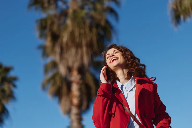 Young woman with curly hair wearing a red coat laughs joyfully while talking on the phone outdoors under a clear blue sky with palm trees in the background.