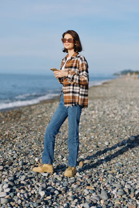 Woman walking on a pebbled beach with a phone, sunglasses, casual plaid shirt, jeans and boots under a sunny coastal sky, capturing a relaxed lifestyle moment for travel and fashion