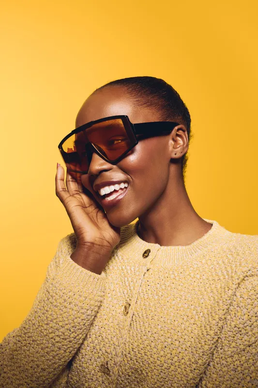 Smiling dark skinned African woman with short hair wearing oversized sunglasses and beige knit sweater poses against yellow background. Concept of fashion, style, beauty, and confidence.