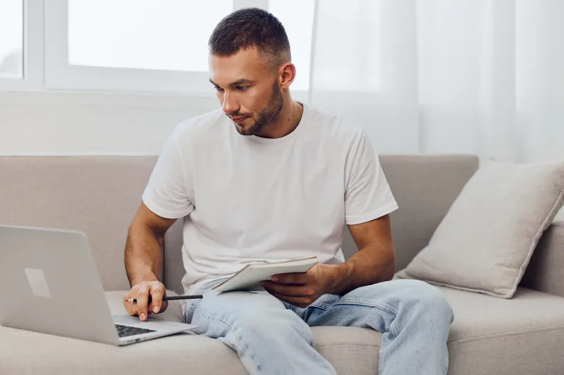 Focused man sitting on a couch, using a laptop and taking notes with a tablet. Casual attire and bright decor create an inviting workspace for productivity.