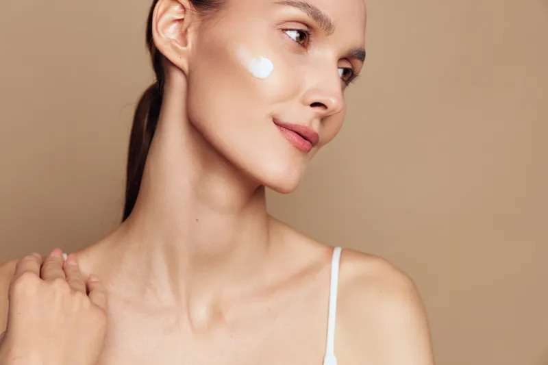 Young woman with clear skin applying white face cream on cheek. Close-up portrait with neutral background. Concept of skincare, beauty, health, and self-care routine.