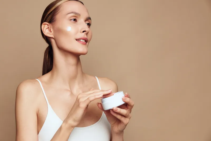 Young woman with long brunette hair applies moisturizer cream on her face while holding a white jar against a plain beige background, expressing calm and self-care.
