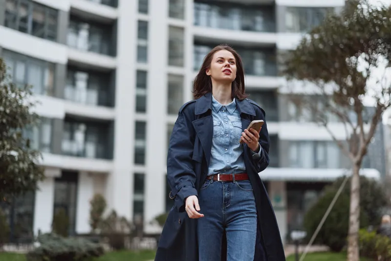 Cityscape scene shows a stylish young woman walking along a sidewalk in denim jeans and shirt under a dark trench coat, projecting confidence and focus against modern buildings.