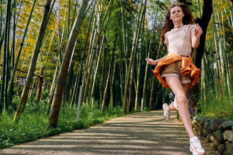 Woman jumping on a gravel path in a bamboo forest, casual outfit with sneakers, joyful happiness and motion captured during a summer walk on a green nature trail with sunlight.