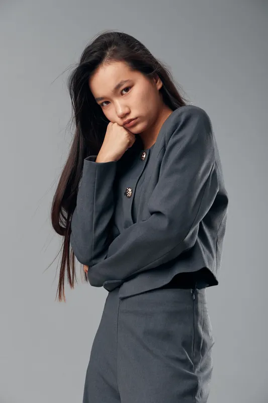 Young woman in gray suit posing with serious expression and thoughtful face standing against gray background with long dark hair and stylish professional outfit.