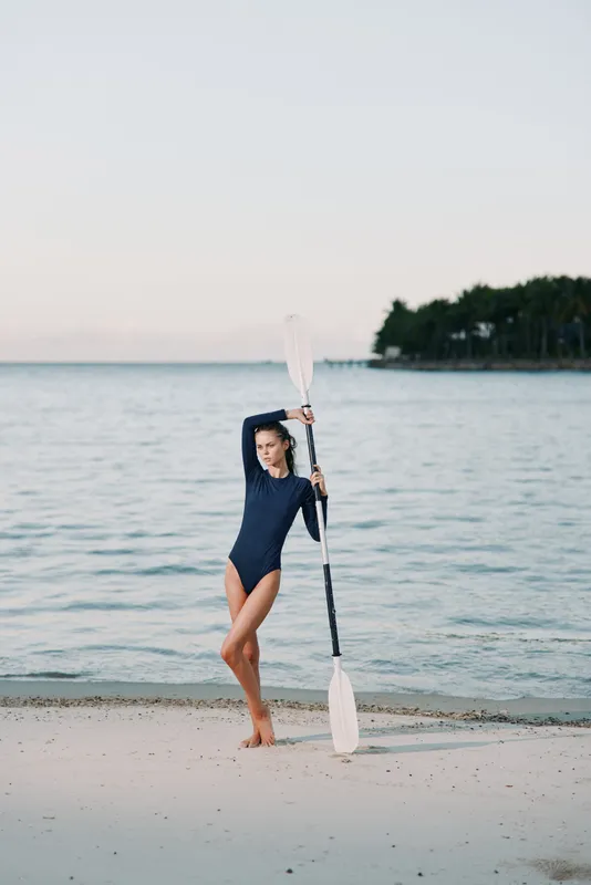 A young woman in a dark swimsuit stands on a sandy beach, lifting a long paddle with one arm as water splashes behind her near the calm shoreline.
