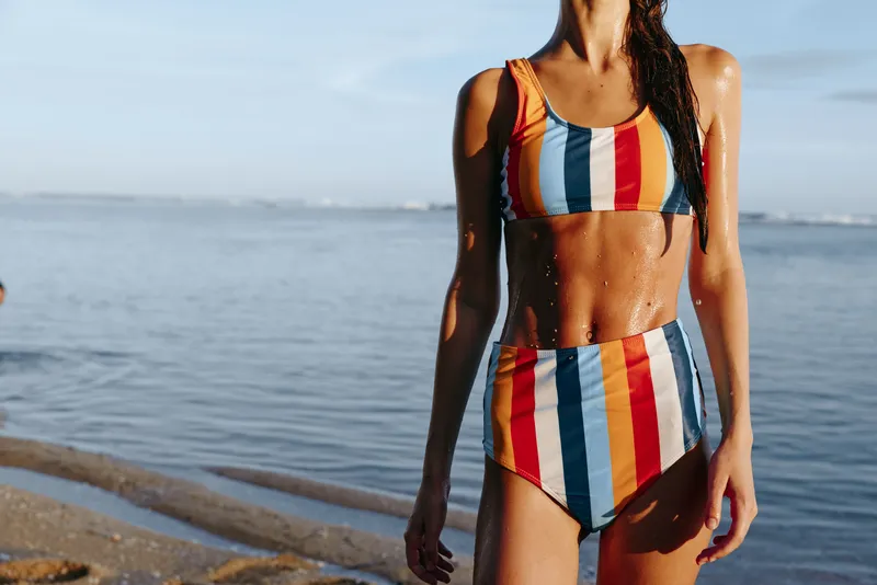 Woman in swimsuit, striped swimwear, stands on beach by ocean, summer day, sunlit skin glistens, outdoors leisure, calm waves, quiet horizon, natural sunlight, vacation