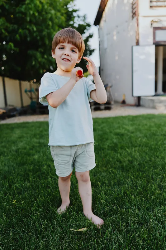 Happy young boy playing outdoors, wearing casual clothing, enjoying the sunshine and green grass, creating a joyful atmosphere in a backyard setting.