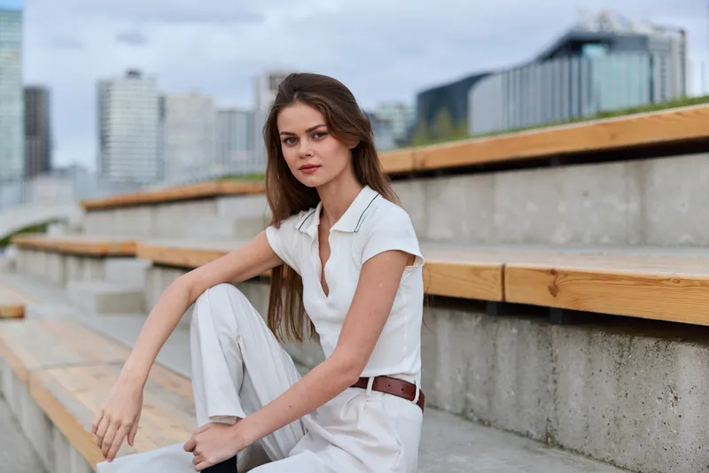 young woman in white outfit sitting on urban stairs with modern cityscape background, casual fashion portrait, natural light, serene expression, soft color palette