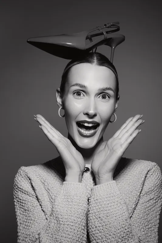 Caucasian woman with fair skin wearing a shoe hat and textured sweater shows surprised expression with hands near face in black and white studio portrait.