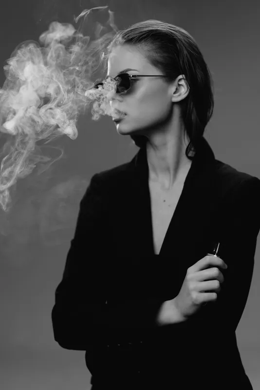 Black and white portrait of a woman in sunglasses exhaling smoke in a dark stylish setting.