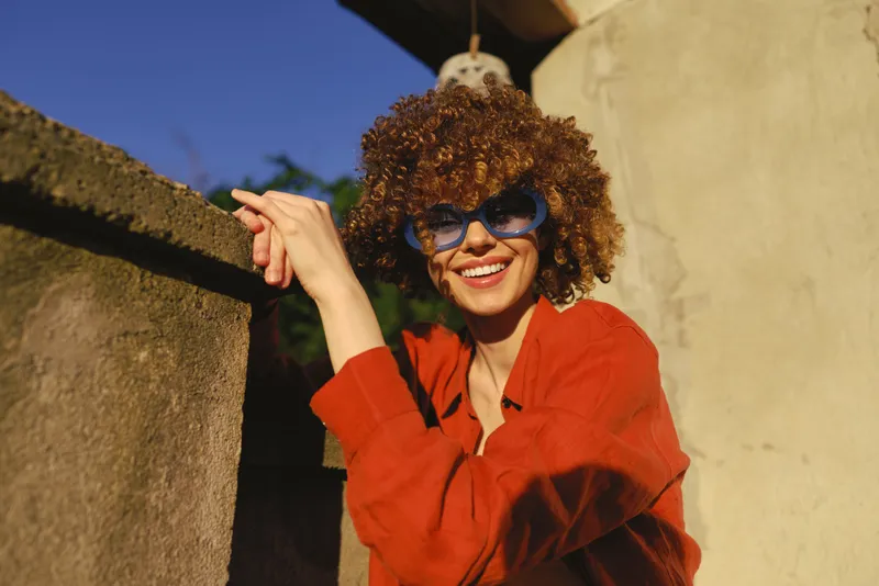 Cheerful woman outdoors, smiling with curly hair, red sweater and sunglasses, sunlit moment, relaxed pose, warm tones, architectural background