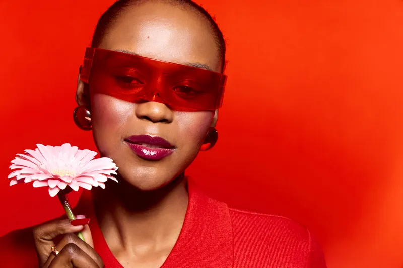 Elegant dark skinned African woman with short hair wearing red sunglasses and red outfit holds pink flower on bright red background. Concept of fashion, beauty, and bold style.
