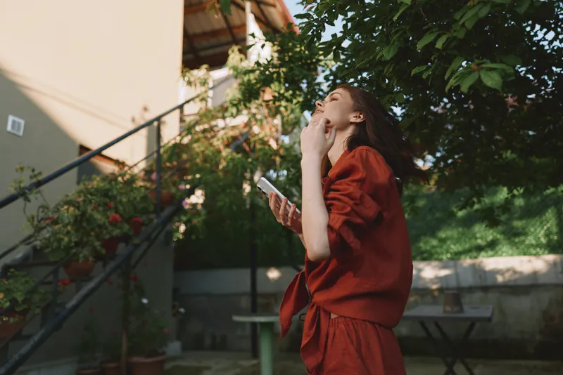 Young woman in a red outfit stands in a sunlit garden, looking upward with a thoughtful expression, one hand raised to her mouth as she holds a small device, capturing a candid moment