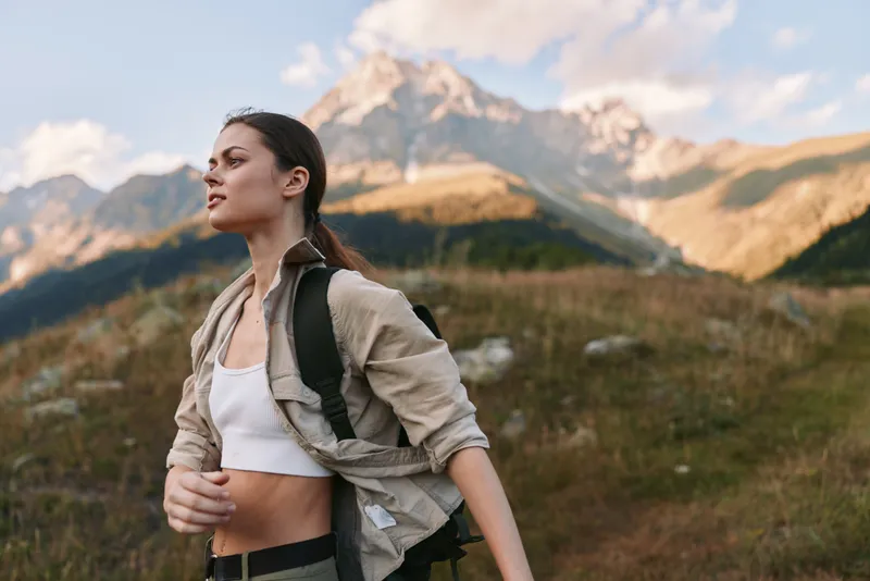Young woman hiking on a mountain trail with a backpack, looking ahead with determination as alpine scenery unfolds behind her, a light jacket flutters in the breeze and her white crop top contrasts
