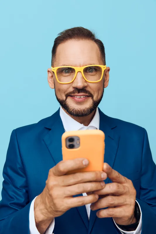 A professional man in a blue suit stands against a solid blue backdrop, looking at a smartphone with a confident, focused face and friendly vibe.